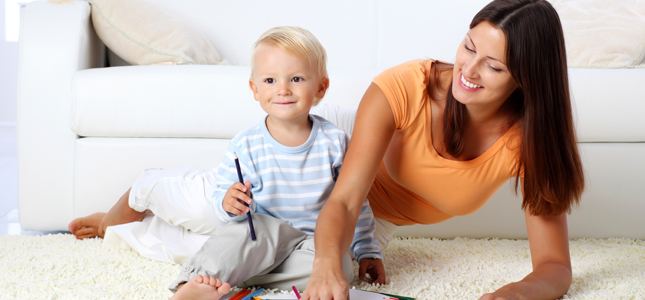 A smiling woman and a young child are coloring together on the floor in a bright living room.
