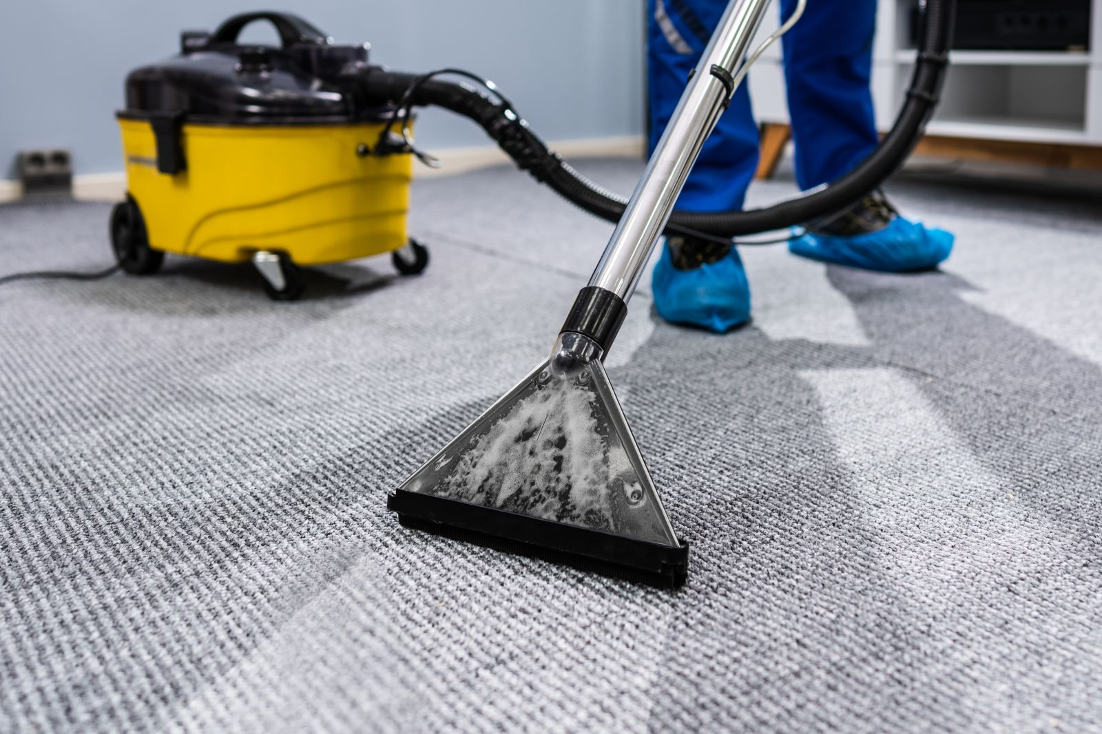 Close-up of a vacuum cleaner being used to clean a grey carpet by a person wearing blue shoe covers.