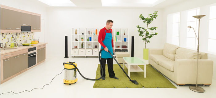 Man vacuuming a green rug in a bright, modern living room with a beige sofa and kitchen area.