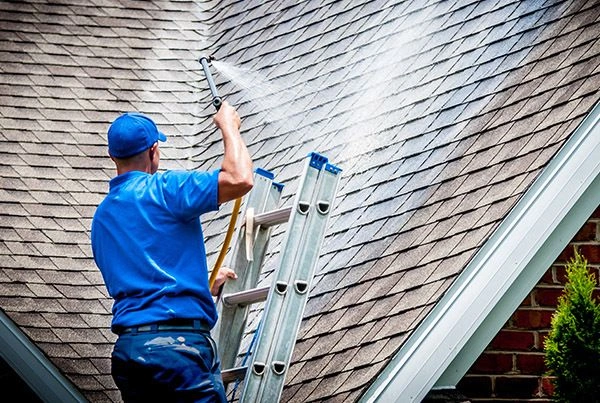 roof-1 Person on a ladder power washing a house roof with water, wearing blue clothes and a cap.